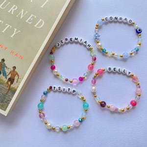 Peut inclure: Quatre bracelets de perles avec des perles de lettres blanches épelant "CONRAD", "JEREMIAH", "BELLY" et "TSITP". Les bracelets présentent des perles colorées dans des tons roses, bleus, verts et des accents dorés. Un livre avec le titre "Summer I Turned Pretty" est en arrière-plan.