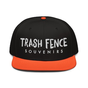 Puede incluir: Gorra de béisbol negra con visera y botón naranja. La gorra presenta el texto blanco "TRASH FENCE" sobre "SOUVENIRS". Un accesorio de moda para un look casual.