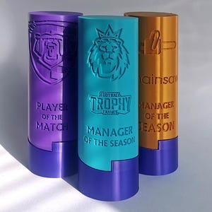 May include: Three cylindrical trophies in purple, teal, and gold. The purple trophy reads "PLAYER OF THE MATCH" with a bear emblem. The teal trophy says "MANAGER OF THE SEASON" with a lion emblem. The gold trophy says "MANAGER OF THE SEASON".