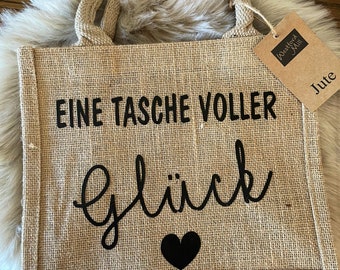 Puede incluir: Una bolsa de mano de yute marrón con asas negras y el texto "Eine Tasche voller Glück" y un corazón negro. La bolsa tiene una etiqueta de papel marrón con el texto "Woolfolk Mill" y "Jute".
