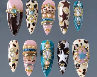 May include: A set of ten long, pointed artificial nails with various designs. The nails feature a mix of colors, including brown, pink, and teal, and are adorned with pearls, gold accents, and star patterns. Some nails have a cow print design.
