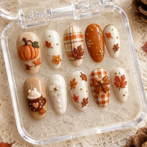 Puede incluir: Un juego de diez uñas postizas en un estuche de plástico transparente, con diseños de temática otoñal que incluyen una calabaza en 3D, una taza marrón con nata montada, patrones de cuadros naranjas y blancos, hojas de arce, flores pequeñas y un lazo marrón.