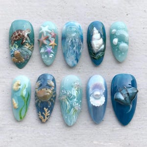 May include: A collection of ten artificial nails with ocean-themed designs. Each nail features a different aquatic creature or element, including a sea turtle, starfish, jellyfish, and seashells. The color palette includes shades of blue, teal, and white.