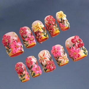 May include: A set of ten artificial nails, each adorned with vibrant floral designs. The nails feature a base of yellow and orange, with pink, red, and white flowers, accented by gold beads and green leaves. The nails are designed for a fashionable manicure.