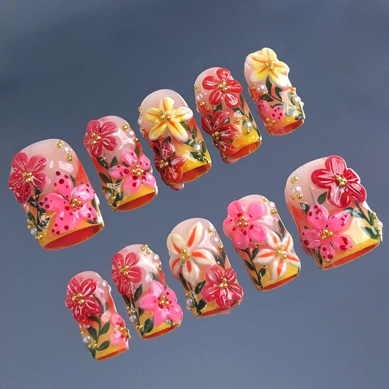 May include: A set of ten artificial nails, each adorned with vibrant floral designs. The nails feature a base of yellow and orange, with pink, red, and white flowers, accented by gold beads and green leaves. The nails are designed for a fashionable manicure.