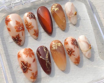 May include: A set of ten almond-shaped press-on nails featuring autumn-inspired designs. The collection includes nails with painted orange maple leaves, solid deep brown tones, gold leaf accents, marble patterns, and thin gold wire embellishments.