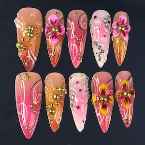 Könnte beinhalten: Eine Sammlung von zehn langen, mandelförmigen Kunstnägeln. Jeder Nagel hat ein einzigartiges Design mit Rosa-, Orange- und Weißtönen, verziert mit goldenen Akzenten, Perlen und 3D-Blumenverzierungen. Die Nägel sind auf einer dunklen Oberfläche angeordnet.