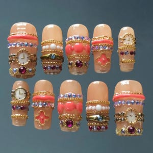 May include: A set of ten artificial nails with a nude base, adorned with gold-colored chains, pink and purple gems, and small clock faces. The nails feature various designs, including beaded accents and decorative shapes.
