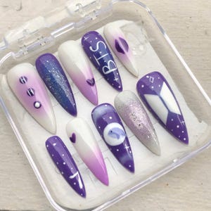 May include: A collection of ten long, almond-shaped false nails in a clear plastic case. The nails showcase diverse designs, including purple gradients, glitter, hearts, and the letters "BTS" in white. Some nails have a white and purple ombre effect.