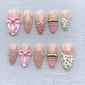 Peut inclure: Un ensemble de dix faux ongles en forme d'amande avec une base nude et divers motifs incluant des nœuds roses en relief, des imprimés léopard dorés, des bandes métalliques dorées et de petits strass transparents sur une surface claire.