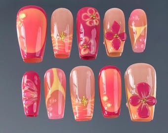 May include: A set of ten artificial nails in various shades of pink, orange, and beige. Some nails feature floral designs, gold accents, and abstract patterns. The nails are arranged on a gray surface.