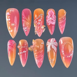 Peut inclure: Dix faux ongles longs en forme d'amande. Chaque ongle présente un dégradé de rose et d'orange, orné d'éléments 3D comme des perles, des papillons et des fleurs. Certains ongles ont des accents clairs en forme de bulles.