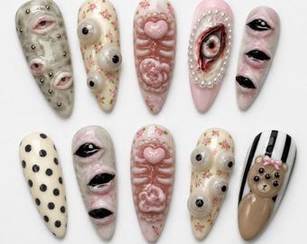 May include: A set of ten press-on nails featuring 3D designs including realistic eyes, floral patterns, pink heart and ribcage motifs, pearl borders, black polka dots, and a brown teddy bear with multiple eyes on a black and white striped background.