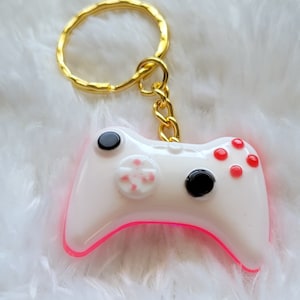 May include: White and pink video game controller keyring with a gold key ring. The controller has red buttons and black accents.
