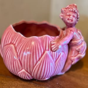 Puede incluir: Un macetero de cerámica rosa con forma de flor de loto con una pequeña figura rosa de una niña que se agarra al lado.