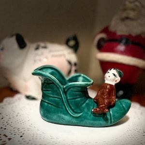 May include: A green ceramic elf shoe with a brown elf sitting on top. The shoe is decorated with a leaf design.