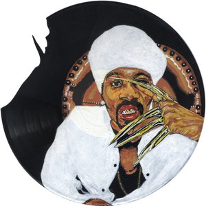 Pode incluir: Um disco de vinil preto pintado com um retrato de uma pessoa usando um chapéu branco e uma camisa branca. A pessoa tem dentes de ouro e unhas longas de ouro. O disco está parcialmente quebrado.