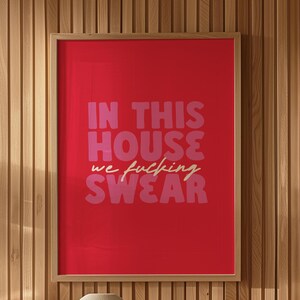 May include: A framed red art print with the text "IN THIS HOUSE we fucking SWEAR" in pink and white. A beige vase sits on a stack of books on a glass table, with a brown chair in the background.