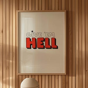 Pode incluir: Uma impressão emoldurada com o texto "GIVE 'EM HELL" em letras vermelhas com contorno preto. A impressão está pendurada em uma parede de painéis de madeira. Uma esfera de cerâmica bege está sobre uma mesa com livros.