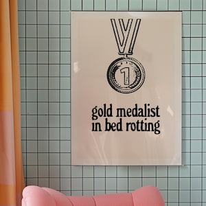 May include: A white poster with a black and white illustration of a gold medal with the number "1" on it. The text "gold medalist in bed rotting" is printed below the medal.