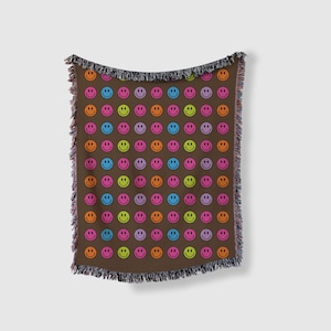 Può includere: Una coperta in tessuto marrone tessuta con un motivo ripetitivo di facce sorridenti colorate. La coperta ha un bordo a frangia.