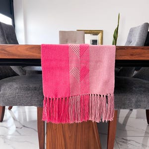 Op de afbeelding: Een roze tafelloper met een geweven ontwerp en franjes. De loper toont verschillende roze tinten, met een centraal gedeelte met een hartpatroon. Een decoratief item voor in huis.