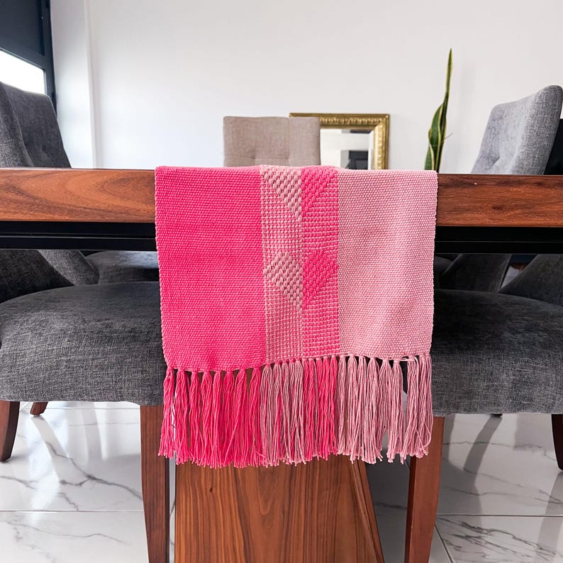 Op de afbeelding: Een roze tafelloper met een geweven ontwerp en franjes. De loper toont verschillende roze tinten, met een centraal gedeelte met een hartpatroon. Een decoratief item voor in huis.