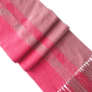 Op de afbeelding: Een roze geweven tafelloper met een geometrisch patroon en gefranjerde randen. De loper toont verschillende roze tinten, waardoor een textiel ontstaat dat geschikt is voor interieurdecoratie.
