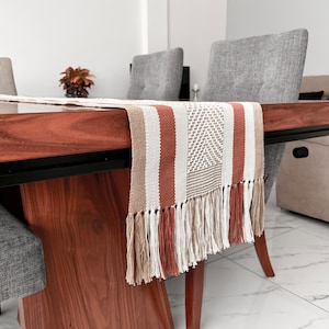 Puede incluir: Camino de mesa tejido con un patrón geométrico y flecos. El camino de mesa presenta rayas verticales en blanco, beige y naranja óxido. La mesa es de madera oscura.