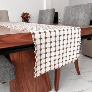 May include: A rectangular, off-white table runner with a repeating brown dot pattern. The runner has a fringed edge and is draped over a dark wood table. The table is set with chairs and a decorative plant.