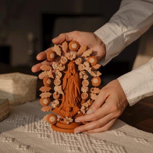 Puede incluir: Una estatua de terracota de una figura religiosa, rodeada por una corona floral. La estatua es sostenida por manos, mostrando los intrincados detalles de las flores y hojas talladas. La figura lleva una larga túnica.