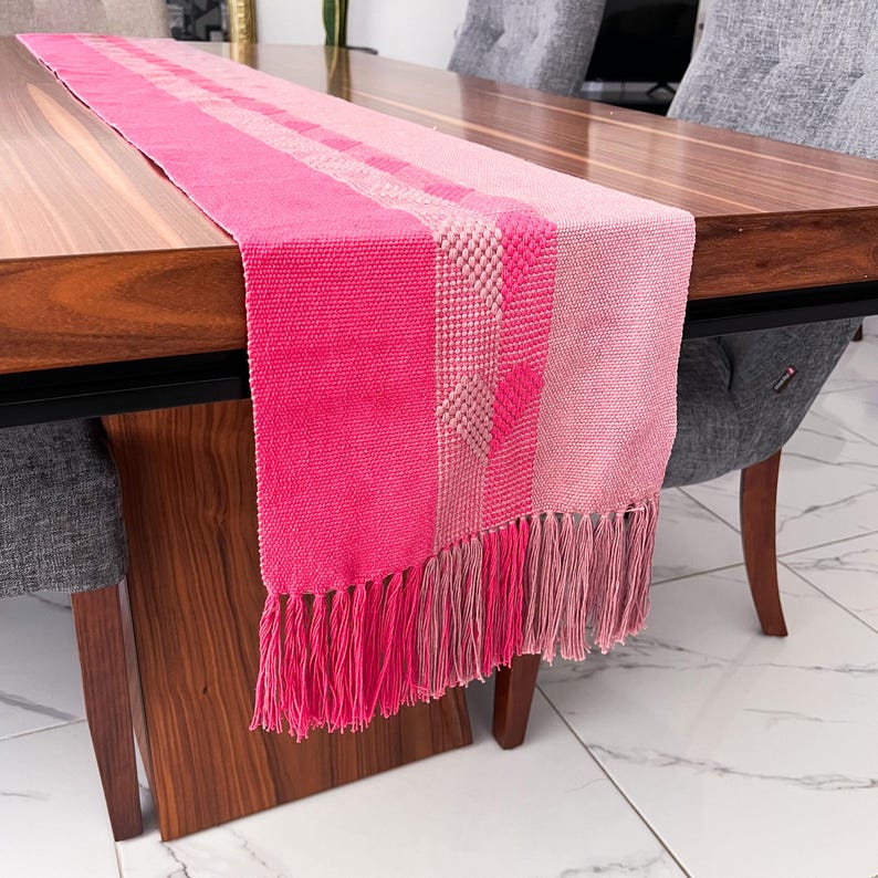 Op de afbeelding: Een roze en fuchsia tafelloper met een gefranjerde rand. De loper heeft een geweven ontwerp met verschillende roze tinten en een textuurpatroon in het midden. Het wordt weergegeven op een houten tafel.