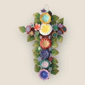 Puede incluir: Una cruz floral artesanal con base marrón y flores coloridas en capas. Las flores son en tonos de amarillo, rojo, azul y morado, con hojas verdes. Un lazo de alambre en la parte superior para colgar.