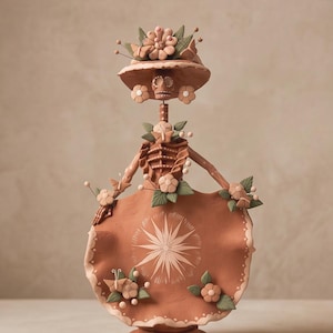 Op de afbeelding: Een terracotta-kleurig kleifiguur van een skelet met een hoed met brede rand, versierd met bloemen en bladeren. Het figuur heeft een skeletachtig bovenlichaam, een lange rok versierd met een centraal sterpatroon en diverse bloemaccenten.
