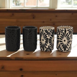 May include: Four ceramic vases. Two are solid black with textured surfaces. The other two are white with a black floral pattern. The vases are cylindrical and sit on a wooden surface.