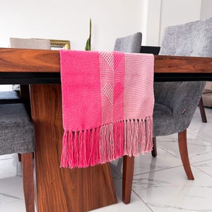 Op de afbeelding: Een felroze tafelloper met een gefranjerde rand. De loper heeft een geweven ontwerp met verschillende roze tinten en een geometrisch patroon. De loper wordt getoond op een donkere houten tafel.