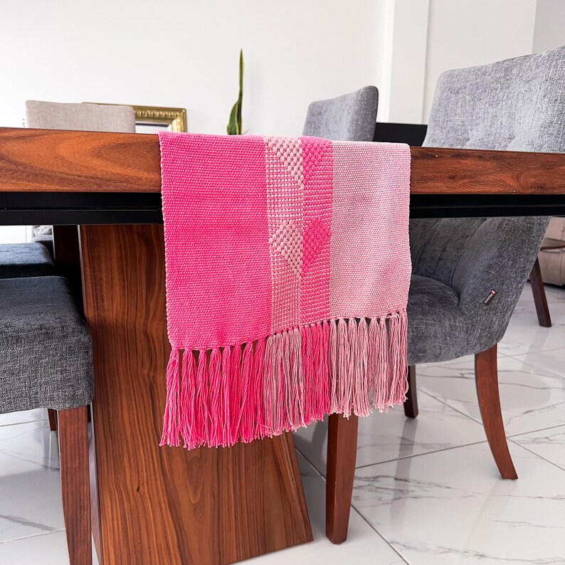 Op de afbeelding: Een felroze tafelloper met een gefranjerde rand. De loper heeft een geweven ontwerp met verschillende roze tinten en een geometrisch patroon. De loper wordt getoond op een donkere houten tafel.