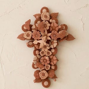 Puede incluir: Una escultura de pared en forma de cruz hecha de arcilla color terracota, con un arreglo intrincado de flores, hojas esculpidas y un colibrí central. La pieza muestra varios tonos de marrón terroso y colores melocotón suaves.