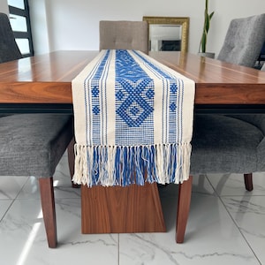 May include: A rectangular table runner featuring a woven pattern in royal blue and off-white. The design includes geometric diamond shapes and vertical stripes, finished with long, dangling fringe tassels in matching blue and white threads.