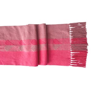 Op de afbeelding: Een geweven sjaal in roze en roze tinten. De sjaal heeft een gestreept patroon met een textuurontwerp en gefranjerde randen. Het kleurenpalet omvat lichte en donkere roze tinten.