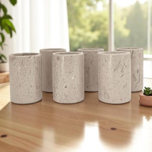 May include: Six cylindrical, speckled gray stone containers are arranged on a light brown wooden table. The containers are of similar height and diameter, with a smooth, polished finish. A small potted plant is visible in the background.