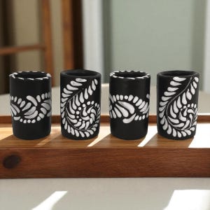 May include: Four black ceramic shot glasses with white floral designs. The glasses are cylindrical and sit on a wooden tray. The white design wraps around the glasses, creating a decorative pattern. The shot glasses are evenly spaced.
