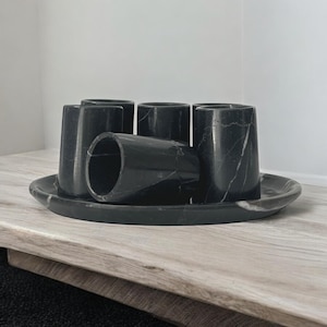 May include: Set of six black marble cups with white veining, arranged on a matching black marble tray. The cups are all the same size and shape, and they are arranged in a circle.