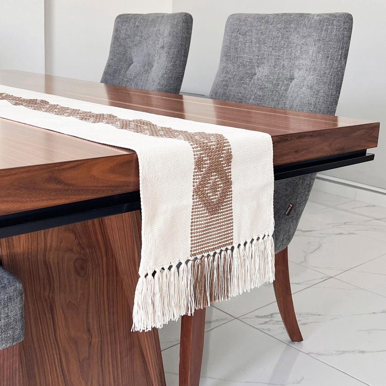 Op de afbeelding: Een cr&egrave;mekleurige tafelloper met een bruin geometrisch patroon en franjes. De loper ligt op een donkere houten tafel, met twee grijze gestoffeerde stoelen op de achtergrond. De tafel is ongeveer 152 cm lang.