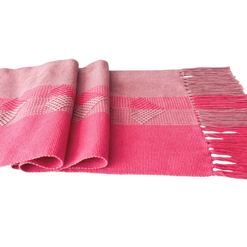 Op de afbeelding: Een geweven tafelloper in roze en roze tinten. De loper heeft een geometrisch patroon en franjes aan &eacute;&eacute;n uiteinde. De stof heeft een getextureerde weefsel, wat visuele interesse toevoegt. De loper is opgevouwen en toont zijn lengte en textuur.
