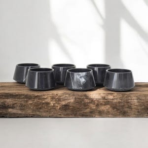 May include: Six black marble cups with a white veining pattern. The cups are arranged in a row on a rustic wooden surface.