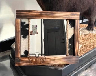 May include: A rectangular mirror framed in dark stained wood with cowhide accents on the left and right sides. Thick tan rope detailing runs vertically along the inner edges of the frame. The brand name Cattle Co Customs is etched on the bottom.