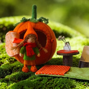 Op de afbeelding: Een gevilt oranje pompoenhuisje met een groene steel, een kleine pop met gevlochten haar in een groene tuniek en oranje muts, een miniatuur houten tafel met een klein glazen vaasje, een houten stoel en een groen-oranje slaapzak met stippen.