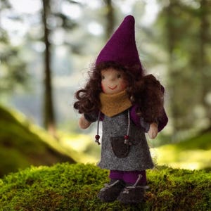 Puede incluir: Muñeca de fieltro hecha a mano con un gorro puntiagudo morado, cabello rizado castaño y un vestido gris. La muñeca tiene una bufanda beige y leggings morados. Se encuentra sobre musgo verde en un bosque.