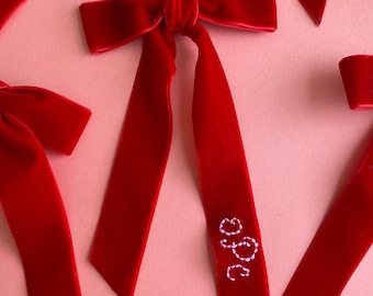 May include: Red velvet ribbon bow with long tails, tied in a knot. The tails are embroidered with the letters "GGG" in white thread.